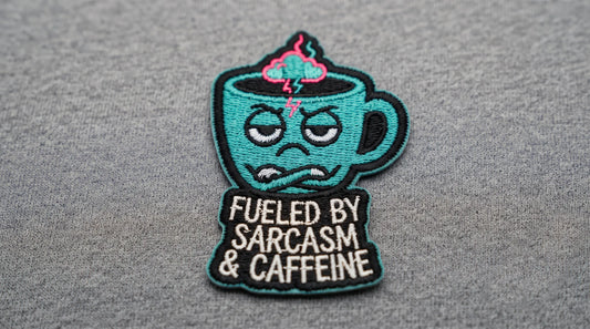 A witty embroidered patch shaped like a coffee cup with eyes, featuring the text "Fueled by Sarcasm & Caffeine."