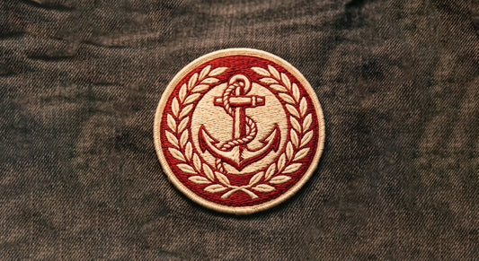 A classic, circular 3D embroidered crest, featuring a fouled anchor encircled by a laurel wreath, centered on a piece of washed black denim. The design is rendered in cream and red threads, facing perfectly forward.
