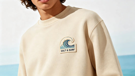 Model wearing clean sand surf hoodie with wave 3D puff embroidery patch on left chest, bright coastal studio background.