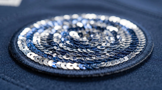 An extreme macro close-up photograph of a single, complete, circular cupped sequin patch, perfectly centered and shown in full circumference on textured blue uniform fabric. The focus is ultra-sharp on the high-stitch density weaving and the flawless, clean-singed edge, illustrating zero-shedding engineering.